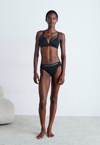 Black bra and matching bikini bottom made of soft fabric, featuring a triangular bra design and logo waistband. Model stands on a light carpet.