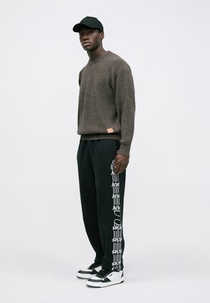 Brown textured sweater with ribbed details; black sweatpants featuring white graphics along the leg; paired with black cap and sneakers.
