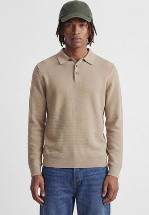 Jeune homme portant un polo en maille beige à manches longues, un jean bleu et une casquette de baseball vert olive, debout devant un fond uni.