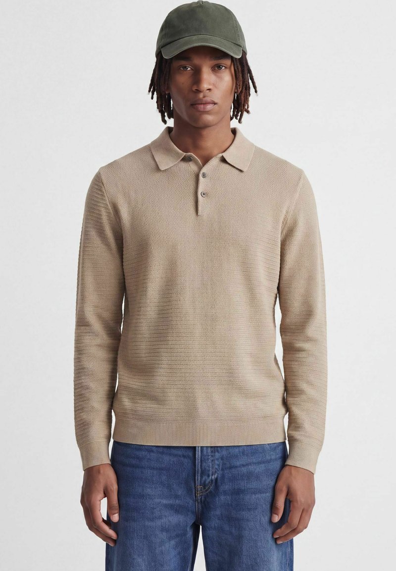Jeune homme portant un polo en maille beige à manches longues, un jean bleu et une casquette de baseball vert olive, debout devant un fond uni.