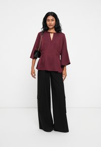 Top bordeaux con scollo a chiave e vita arricciata, abbinato a pantaloni neri a gamba larga con tasche cargo.