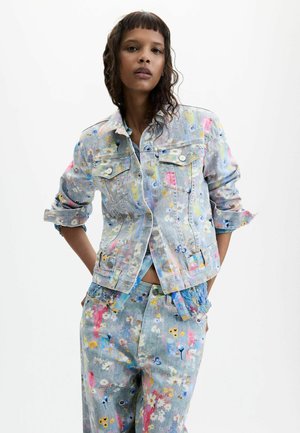 Frau trägt hellblaue Jeansjacke und Hose mit buntem floralen Farbspritzmuster, steht mit den Händen in den Taschen vor weißem Hintergrund.