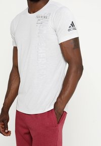 Vit kortärmad sporttröja med rund hals, med svart text och en Adidas-logotyp på vänster ärm. Lättviktig tygstruktur.