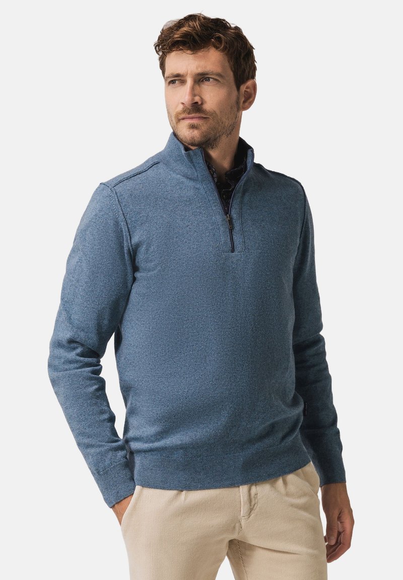 Blue zip-neck sweater in textured knit, featuring a stand collar and ribbed cuffs. Worn over a patterned shirt with beige pants.