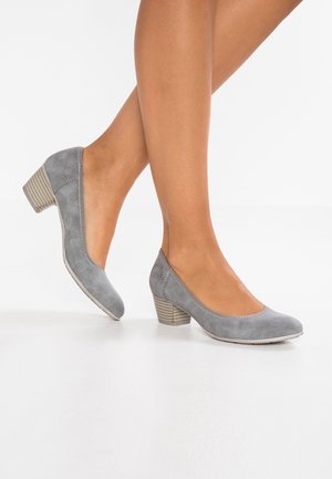 Pies con zapatos bajos de ante gris con punteras redondeadas, de pie sobre una superficie blanca con un fondo liso.