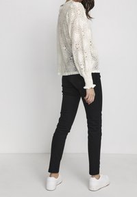Top en dentelle à manches longues blanc cassé, orné de motifs floraux, avec des détails de poignets élastiques, associé à un pantalon noir slim et des baskets blanches.
