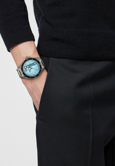 Isik, kes kannab musta kampsunit ja pükse, vasak käsi taskus, randmel hõbedane kell hele sinise sihverplaadi ja musta raami ümber.