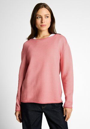 Frau trägt einen rosa-weiß gestreiften Langarmpullover über einem weißen Oberteil, kombiniert mit dunkelblauen Jeans, und steht vor einem weißen Hintergrund.