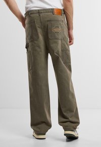Olijfgroene cargo broeken met meerdere zakken, versterkte stiksels en een leren label met het "Another" logo op de achterkant van de tailleband.