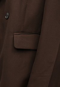 Κοντινό πλάνο ενός καφέ μπουφάν με τσέπη με καπάκι και ένα εμφανές μαύρο κουμπί στην μπροστινή αριστερή πλευρά.