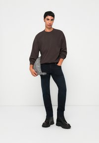 Young man in dark sweatshirt, black jeans, and black boots holding a gray backpack against a white background.