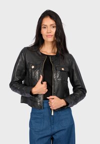 Černá kožená krátká bunda s kulatým výstřihem, dvěma předními kapsami se zlatými detaily a zipovým zapínáním. Nositá přes černou košili.