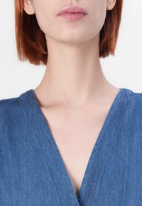 Denim vaate, jossa on V-muotoinen kaula-aukko, joka taipuu sileäksi pinnaksi ja siniseen sävyyn. Suunnitelma sisältää ommeltua yksityiskohtaa reunoilla.