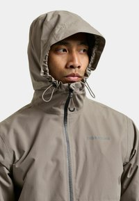 Chaqueta impermeable beige con capucha ajustada alrededor del rostro, con cremallera frontal y un logotipo en el pecho. Textura lisa.