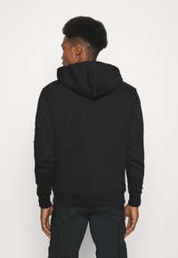 Sweatshirt à capuche noire avec une poche kangourou, des poignets et un ourlet côtelés. Présente un tissu texturé et une poche zippée sur la manche.