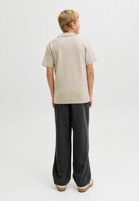 Polo en maille beige à manches courtes, associé à un pantalon ample gris foncé et des chaussures à enfiler couleur beige, vu de dos.