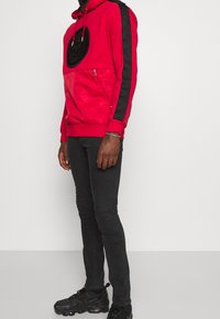 Man i röd huvtröja med svarta vertikala ränder på ärmarna, svarta smala jeans och svarta sneakers, stående mot en enkel vit bakgrund.