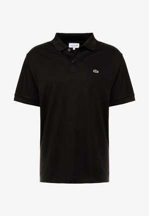 Person trägt schwarzes Polo über weißem und grün gestreiftem Langarmshirt, weiße Hose und schwarze Sneaker mit weißen Sohlen.