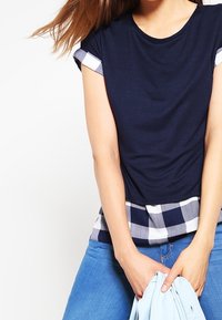 Mörkblå t-shirt med korta ärmar och vita detaljer, med ett rutigt nederkant i mörkblått och vitt. Bärs över ljusblå jeans.