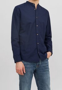 Man som bär en marinblå skjorta med mandarinkrage, knäppt med långa ärmar och blå jeans, med ena handen i fickan mot en enfärgad bakgrund.