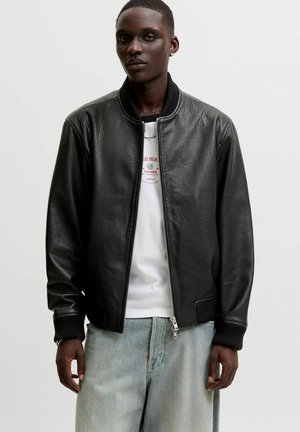 Jeune homme portant une veste bomber en cuir noir sur un t-shirt blanc à motif graphique et un jean bleu clair, debout devant un fond uni.