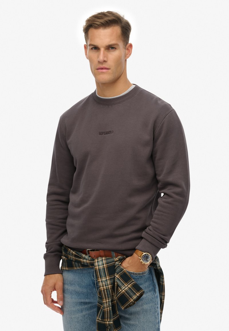 Szara bluza z okrągłym dekoltem i długimi rękawami, z subtelnym logo na piersi. Połączona z niebieskimi dżinsami i koszulą w kratę zawiązaną w talii.