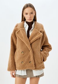 Giacca in pelliccia sintetica marrone e soffice con design doppiopetto, colletto ampio e due tasche frontali. Indossata sopra un dolcevita marrone e una gonna a scacchi.