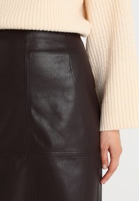 Jupe en cuir marron avec taille haute, texture lisse et coutures visibles. Accompagnée d'un pull en maille de couleur crème.