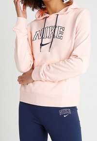 Person som bär en ljusrosa Nike-hoodie med marinblå dragsnören och marinblå leggings, båda med Nike-logotyp och text.