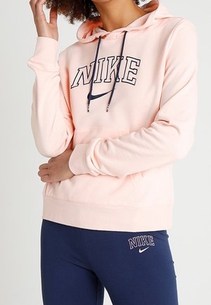 Persona vestida con una sudadera con capucha rosa claro de Nike con cordones azul marino y leggings azul marino, ambos con el logo de Nike y texto.