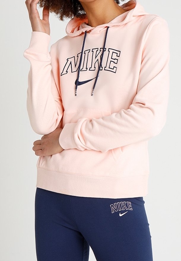 Person som bär en ljusrosa Nike-hoodie med marinblå dragsnören och marinblå leggings, båda med Nike-logotyp och text.