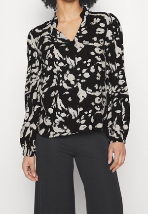 Kvinde iført en sort og hvid bluse med abstrakt print, lange ærmer og V-hals samt sorte bukser med vide ben.