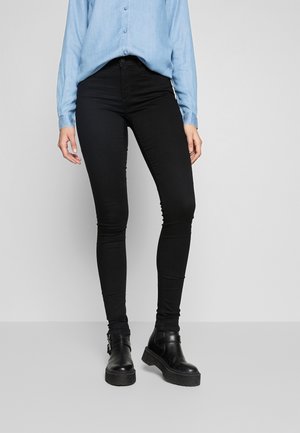 Femme portant un jean skinny noir, des bottines noires, et une chemise à manches longues bleu clair, se tenant devant un fond neutre.