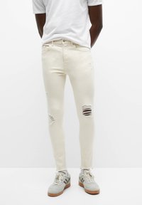 Jeans blancos ajustados con desgastes y rasgaduras en las rodillas, combinados con zapatillas grises. El material parece denim con una textura suave.