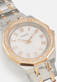 Montre octogonale avec bracelet en acier inoxydable, accents en or rose, cadran texturé blanc, index en chiffres romains et lunette ornée de pierres scintillantes.