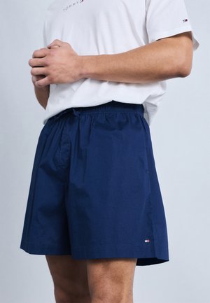 Mann iført marineblå shorts og en hvit Tommy Hilfiger t-skjorte, stående med hendene sammenfoldet foran mot en ensfarget bakgrunn.