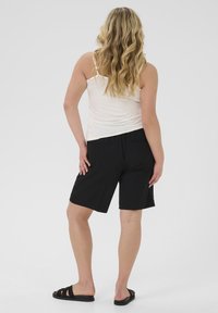 Pantaloni corti neri con una vestibilità rilassata, due tasche posteriori, abbinati a una canotta bianca. Il tessuto appare liscio e leggero. Sandali completano il look.