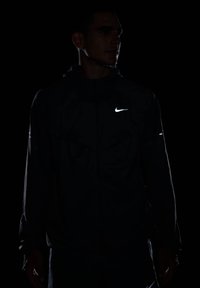 Giacca nera Nike con logo e accenti riflettenti, dotata di cappuccio, chiusura a zip e tessuto testurizzato, indossata da un modello in un ambiente buio.