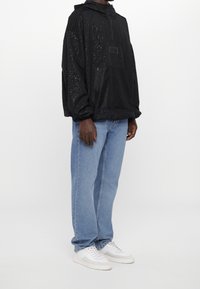 Person som bär en svart prickig huva-jacka, ljusblå raka jeans och vita sneakers som står mot en neutral bakgrund.