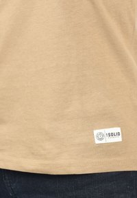 Beige bomulds t-shirt med en glat tekstur, der har et hvidt mærke syet i kanten, som viser mærkenavnet "!SOLID" og logo.