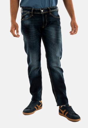 Jeans in denim blu scuro con dettagli scoloriti, design classico a cinque tasche e cuciture a contrasto. Abbinati a sneakers nere con accenti arancioni.