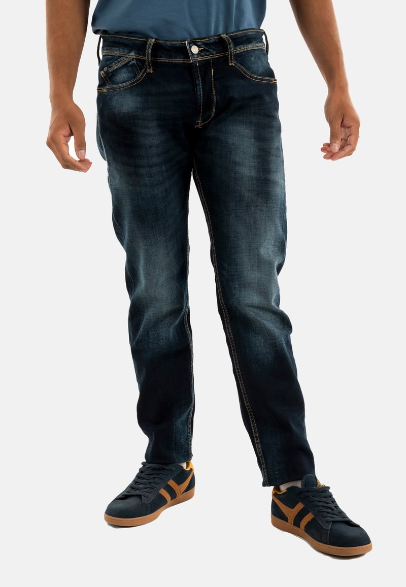 Jeans de mezclilla azul oscuro con detalles desgastados, diseño clásico de cinco bolsillos y costuras en contraste. Combinados con zapatillas negras con acentos naranjas.