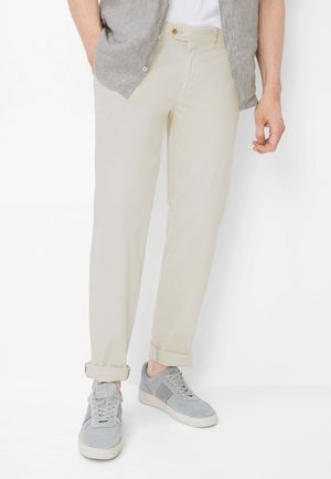 Man met beige opgerolde broek, grijze sneakers, wit overhemd en lichtgrijs open knoopoverhemd, staand tegen een witte achtergrond.