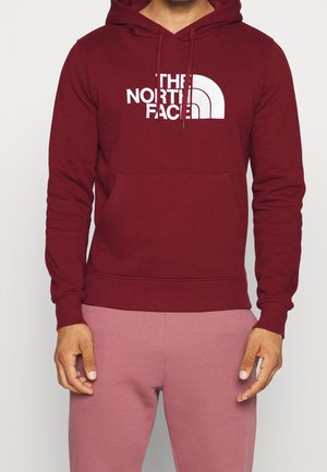 Mężczyzna w bordowej bluzie z kapturem z białym logo "The North Face" i liliowymi spodniami dresowymi, stojący na jednolitym tle.