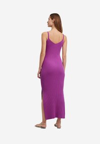 Lila gerippter Strickkleid mit dünnen Trägern, tiefem Rückenausschnitt, figurbetonter Form und Seitenschlitzen am Saum. Sandalen sind an den Füßen sichtbar.