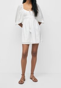 Weißes Leinenkleid mit Puffärmeln, quadratischem Ausschnitt, Taillenbund mit Kordelzug und Quastenakzenten. Kombiniert mit braunen Sandalen mit Riemchen.
