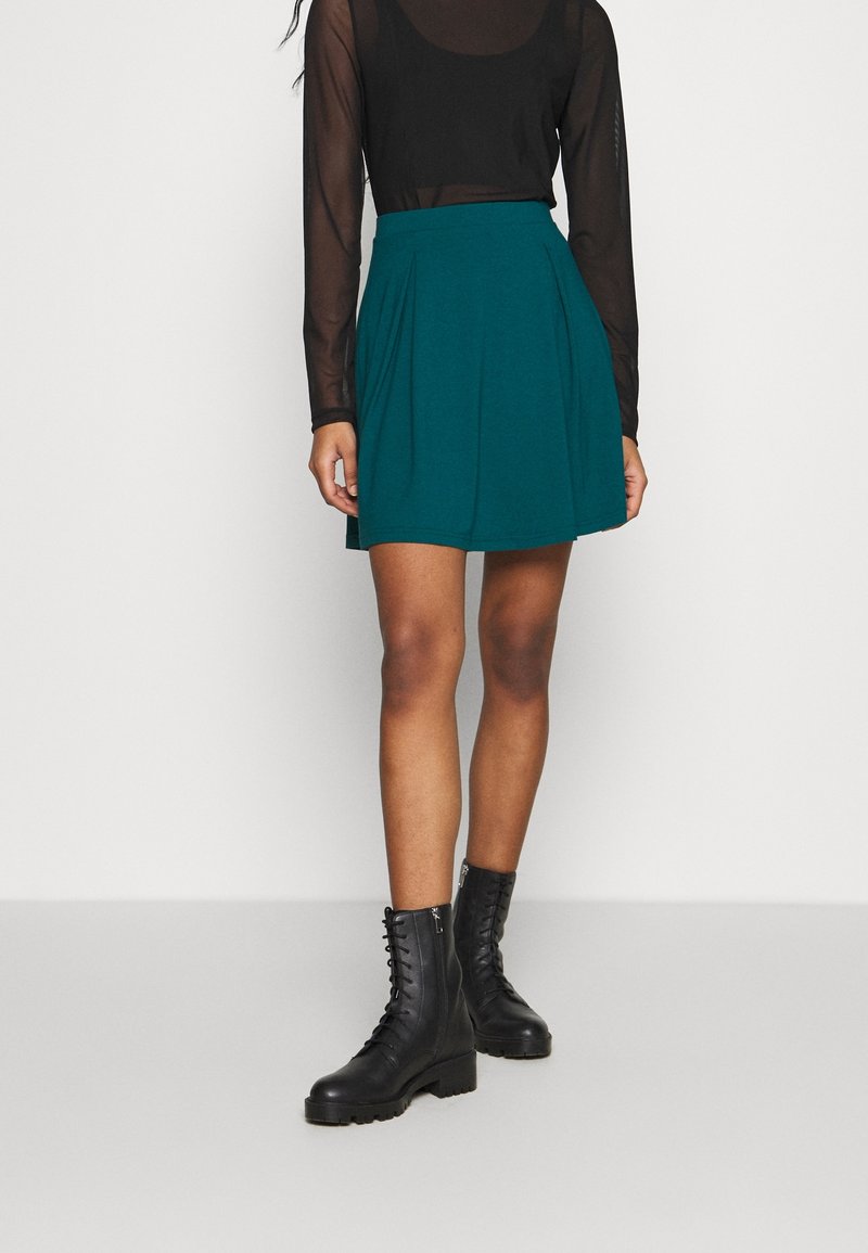 Personne portant un haut noir transparent à manches longues, une jupe plissée turquoise et des bottes de combat noires lacées, debout devant un fond uni.