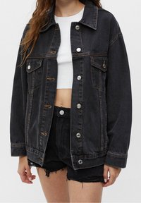 Giacca in denim nera con taglio classico, tasche frontali, bottoni argentati e cuciture a contrasto. Indossata sopra una maglietta crop bianca e pantaloni shorts neri.