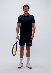 Νεαρός άνδρας με μαύρη αθλητική φόρμα που κρατά ρακέτα τένις, στέκεται μπροστά σε απλό λευκό φόντο.