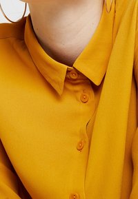 Senfgelbe Bluse mit klassischem Kragen, ausgestattet mit orangen Knöpfen und einer glatten, weichen Stoffstruktur. Modisches und zeitgemäßes Design.